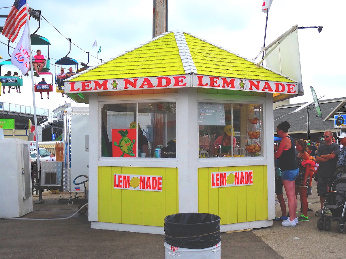 Puesto de limonada en una feria. El puesto tiene un techo amarillo brillante y paneles laterales. La palabra “Limonada” está escrita con letras rosadas brillantes en la parte de arriba y en los lados.