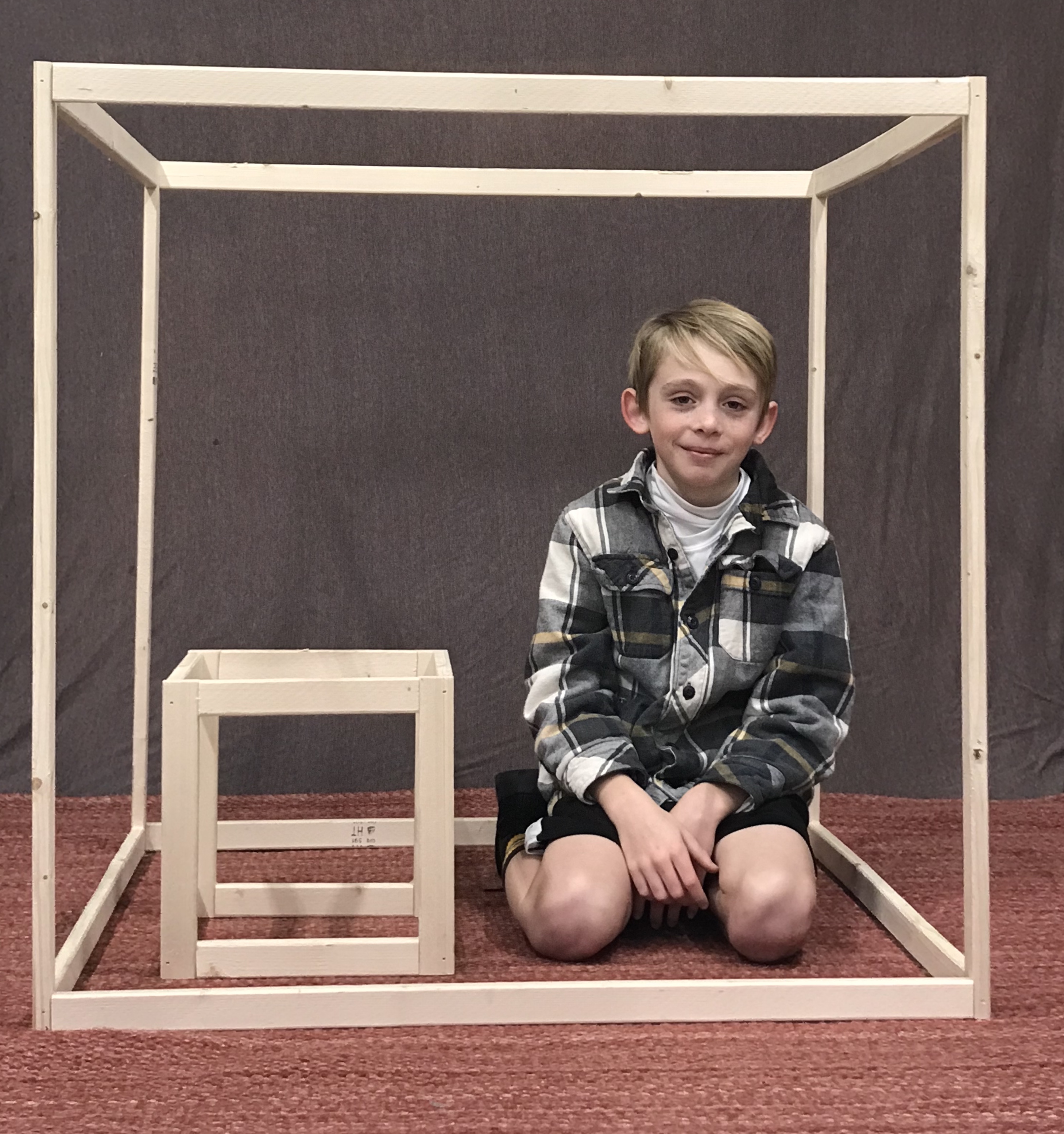A student and two rectangular prisms. . The student is sitting inside the large rectangular prism and is next to the smaller prism. 