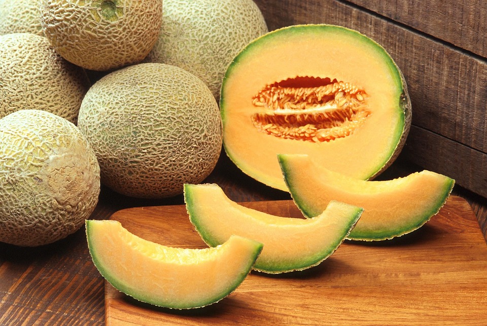 Varios melones cantalupos en una mesa. Un melón está partido por la mitad y hay tres pedazos de melón en una tabla de cortar. Los pedazos son anaranjados y tienen cáscara verde.
