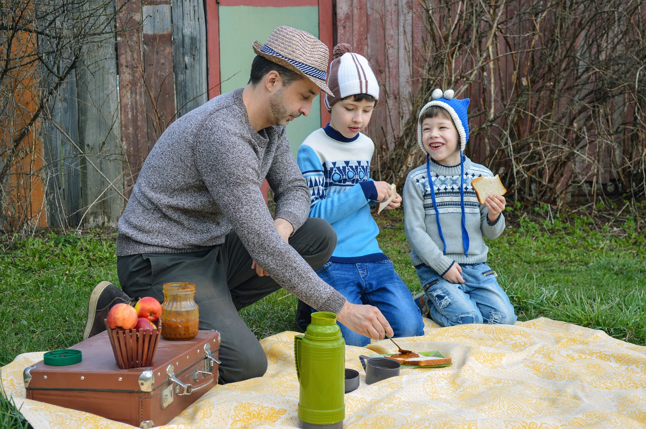 PIcnic outside. 3 people. 1 man, 2 boys. Making a sandwich. Eating a sandwich. Apples in a basket.