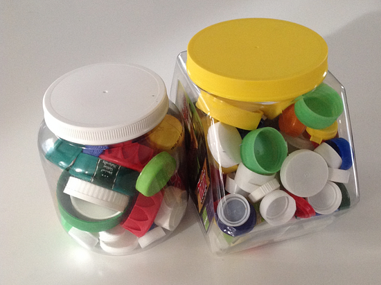 A photo of two clear, plastic containers containing plastic bottle caps of various sizes and colors. The colors of the plastic bottle caps are red, orange, yellow, green, blue, and white.