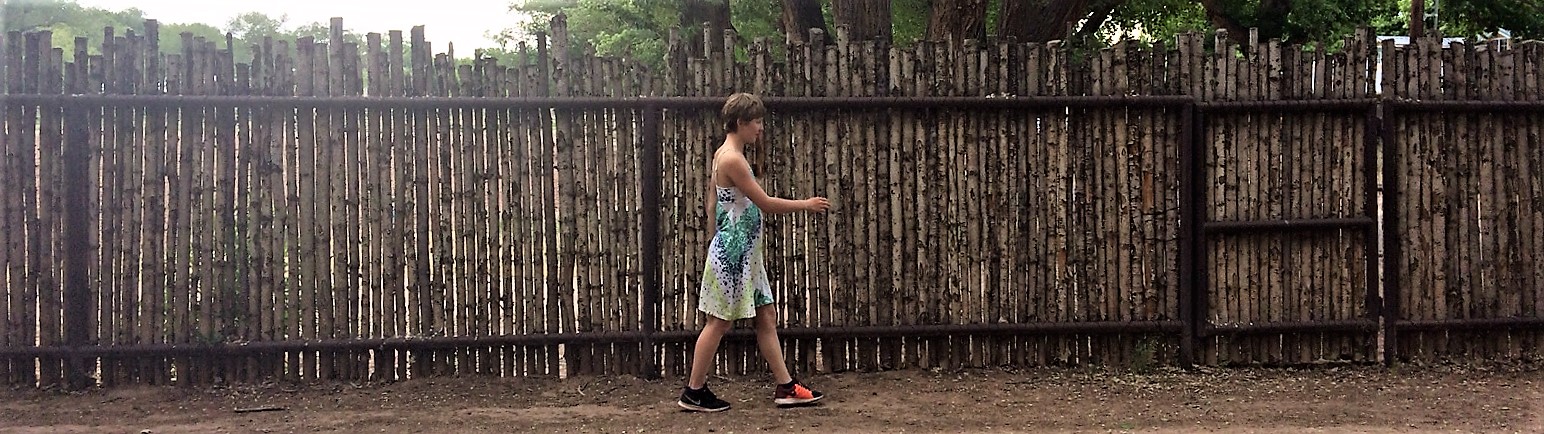 A photo of a woman walking along a wodoen fence.