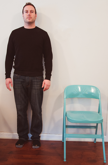 A picture of a man standing next to a folding chair. The top of the folding chair comes up to the man's hip.