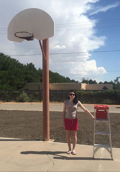 An image of a full size basketball hoop attached to a pole. To the right of the pole stands a young woman. The height of the woman stops about midway up the pole. To the right of the woman stands a 4-rung step ladder. The height of the step ladder stops at the woman’s shoulder.